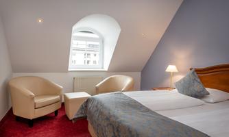 A cozy bedroom with a double bed and two comfortable armchairs. The room has a sloping skylight and warm light.