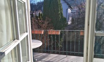 A sunny balcony with a table and a view of green trees. Plant boxes are visible behind the railing.