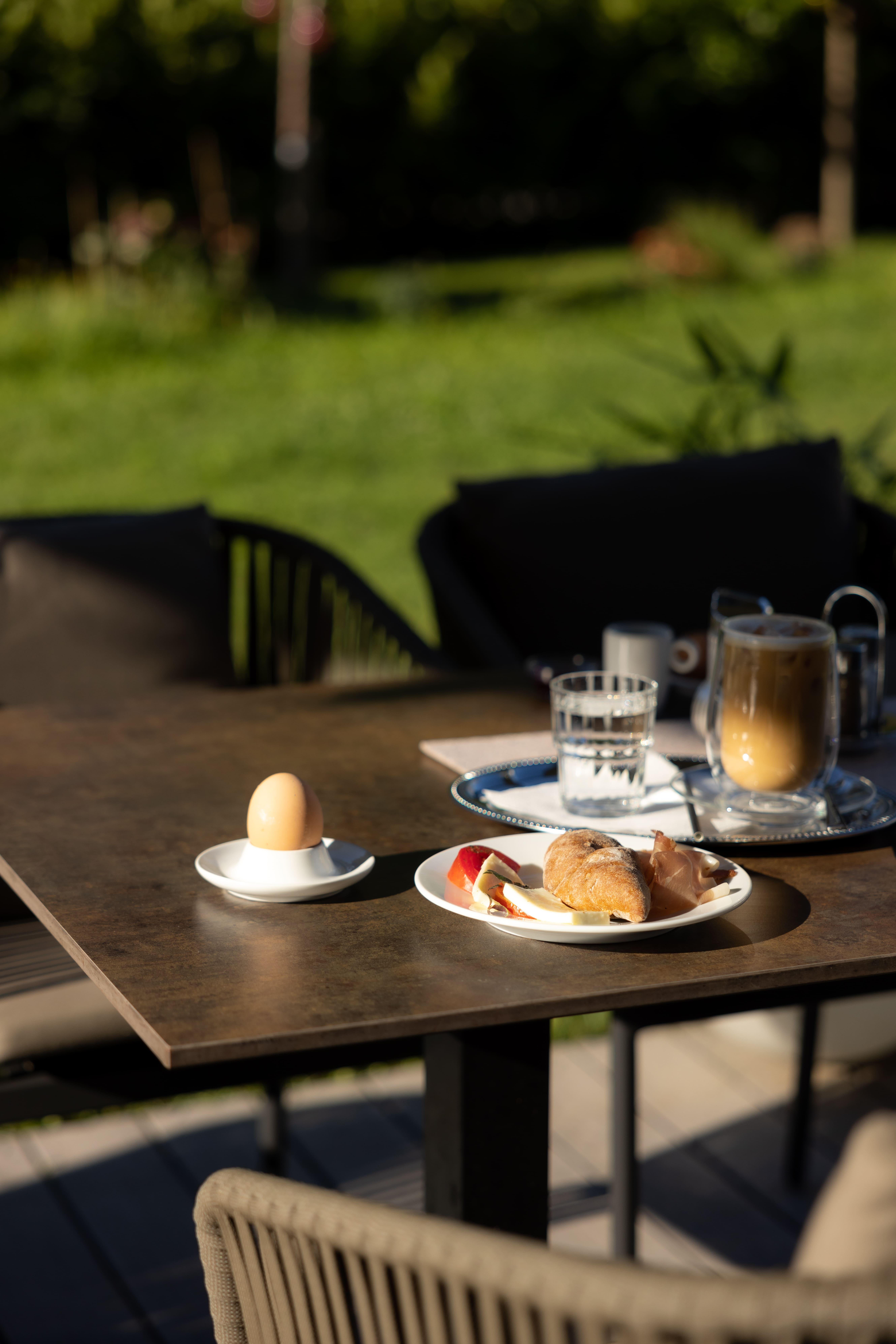 A beautifully set table outdoors with a breakfast set. There is an egg, a croissant, apple slices, and drinks.