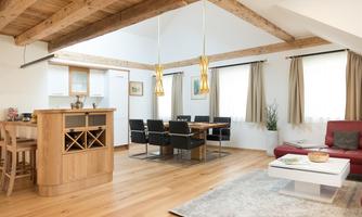 A modern living room with an open kitchen area. The room has wooden beams, light furniture, and large windows that provide plenty of light.