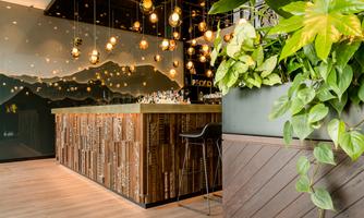 A stylish restaurant with a wooden bar and modern pendant lights. Large plants create a green and inviting atmosphere.