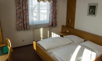 A simple bedroom with two beds and natural light. There is a small seating area and curtains with floral patterns.