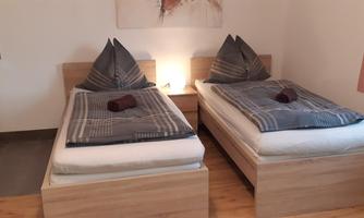A simple room with two single beds. The beds are neatly made and there is a small kitchenette in the background.