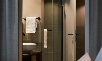 A modern bathroom with a glass shower and a sink. In the foreground, there is a cozy bedroom with bedding visible.