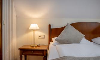 A cozy bedroom with a large bed and a small bedside lamp. The bedding is white and the pillows are arranged decoratively.