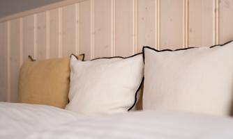 A cozy bed with several pillows in soft colors. The wooden wall gives the room a warm atmosphere.