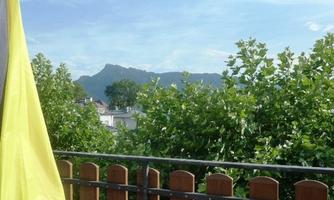 A terrace with a wooden railing and a view of green trees. In the background, mountains and a blue sky can be seen.