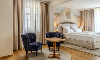 An elegant room with a large bed and two blue chairs at a small table. The windows are decorated with light curtains and the wooden floor radiates warmth.