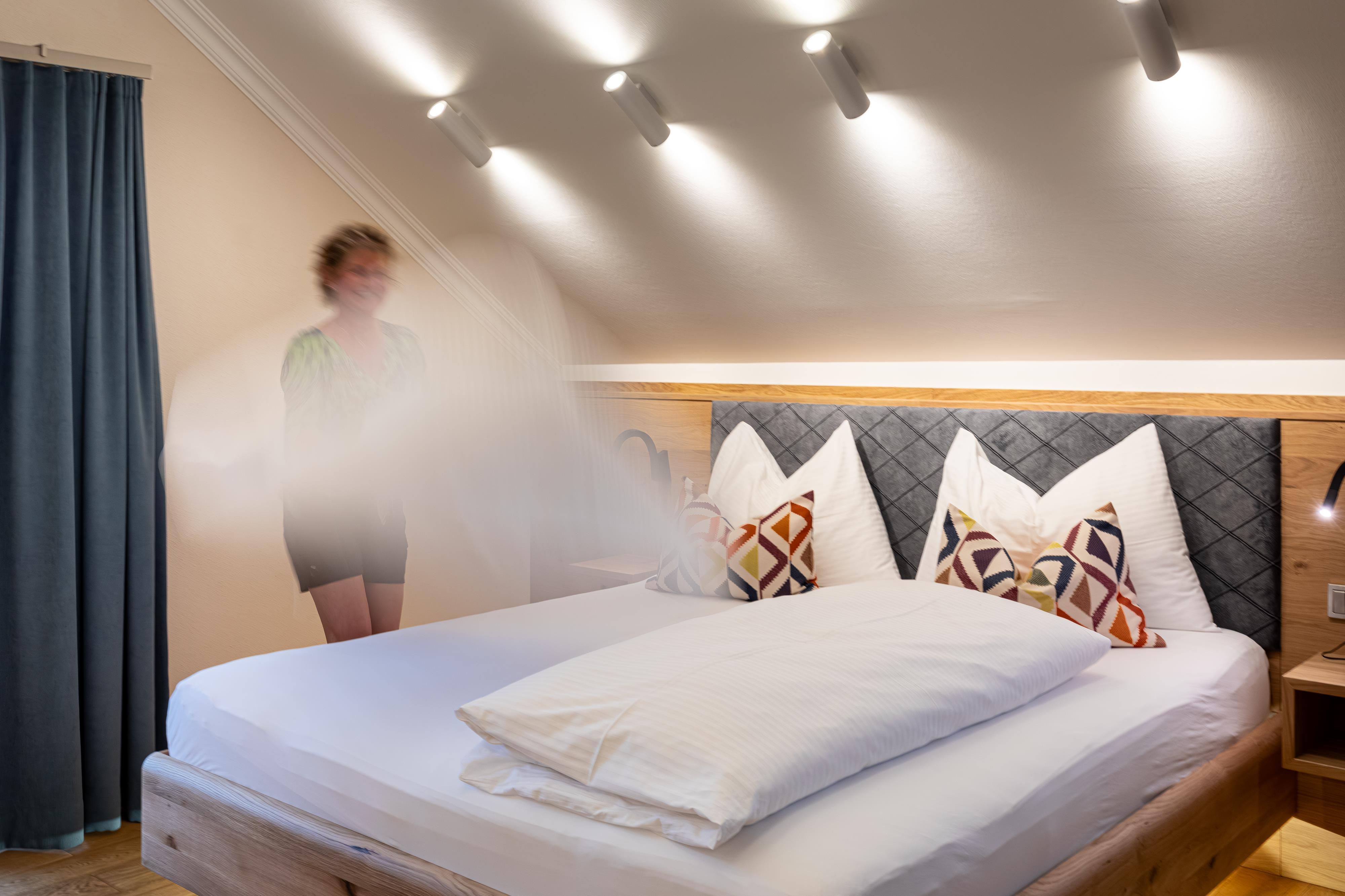 A cozy bedroom with a large bed and colorful pillows. In the background, there is a person holding bed linens.