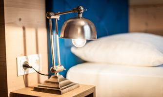 A modern table lamp stands on a nightstand next to a cozy bed. The background is in a calming blue.