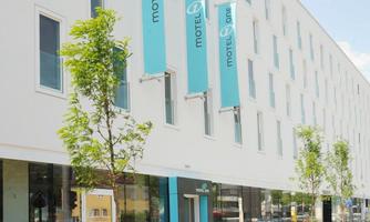 A modern hotel building with a bright facade and large windows. In front of the hotel, there are small trees and friendly signage is visible.