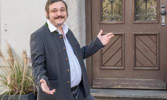 A man with a mustache stands in front of a wooden door and spreads his arms. He is wearing a traditional jacket and smiling kindly.