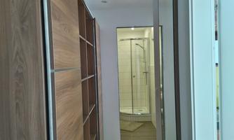 A modern hallway with wooden shelves and a window overlooking the shower cabin. The walls are light and the lighting is soft.