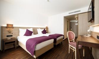 A modern hotel room with two single beds and elegant furniture. The wooden floors and soft lighting create a cozy atmosphere.