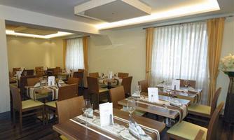 A modern restaurant with wooden tables and chairs. The walls are brightly decorated, and large windows provide plenty of daylight.