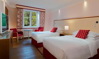 A modern hotel room with two single beds and a small table. Large windows let in a lot of light and offer a view of nature.
