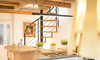 A modern kitchen with a wooden island and an elegant spiral staircase. Green plants adorn the space, and a piece of artwork hangs on the wall.