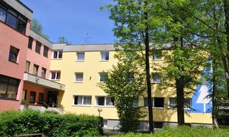 A building with colorful facades in yellow and pink, surrounded by green trees. The sky is clear and blue.