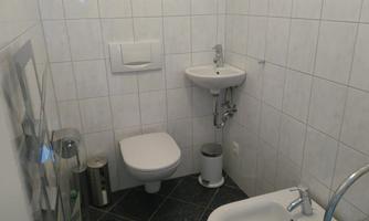 A small bathroom with white tiles. It contains a toilet, a sink, and a waste basket.