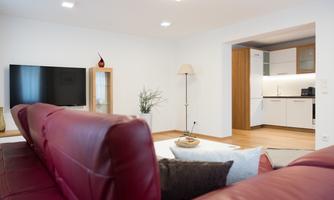 A modern living room with a red sofa and a large television. In the background, a bright kitchen is visible.