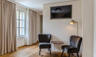 A modern room with two black armchairs, a table, and a TV on the wall. Large windows with curtains let in plenty of light.