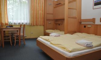 A simple hotel room with a double bed and two single beds. Water bottles are on the table and the windows have bright curtains.