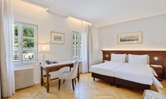 An elegant room with two single beds and a desk. Large windows let in plenty of light and there is a view of the surroundings.