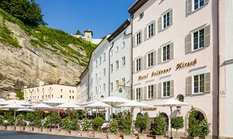 A picturesque hotel building with the inscription "Hotel Goldener Hirsch". In front of the hotel are elegant terraces with sun umbrellas and beautiful plants.