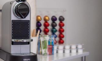 An elegant coffee machine by Krups sits on a table, surrounded by colorful coffee capsules. In the background, various utensils and a king's table are visible.