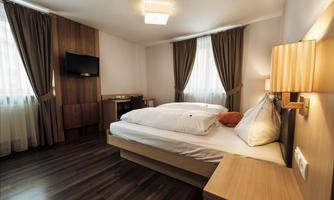 A modern hotel room with a double bed, wooden floor, and large windows. The walls are in neutral colors, and a TV is mounted on the wall.