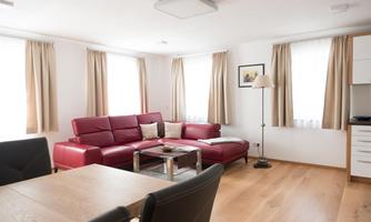 A cozy living room with a red sofa and a wooden table. Bright curtains let in plenty of light.