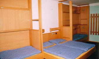A simple dormitory with several beds and wooden shelves. The mattresses have striped covers and there is ample storage space.