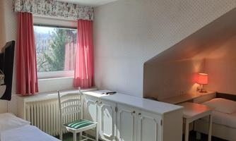 A simple room with two beds, a table, and a chair. It has a window with curtains and soft lighting.