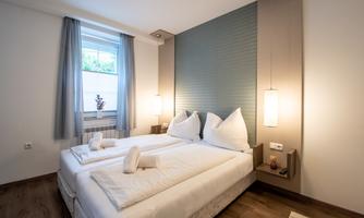 A modern bedroom with two beds and white bedcovers. Large windows and soft lighting create a cozy atmosphere.
