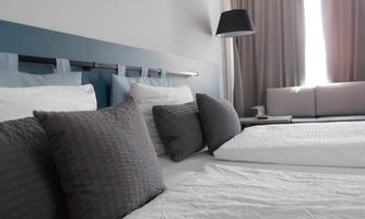 A modern hotel room with two beds and gray pillows. The room has light curtains and an elegant lamp.