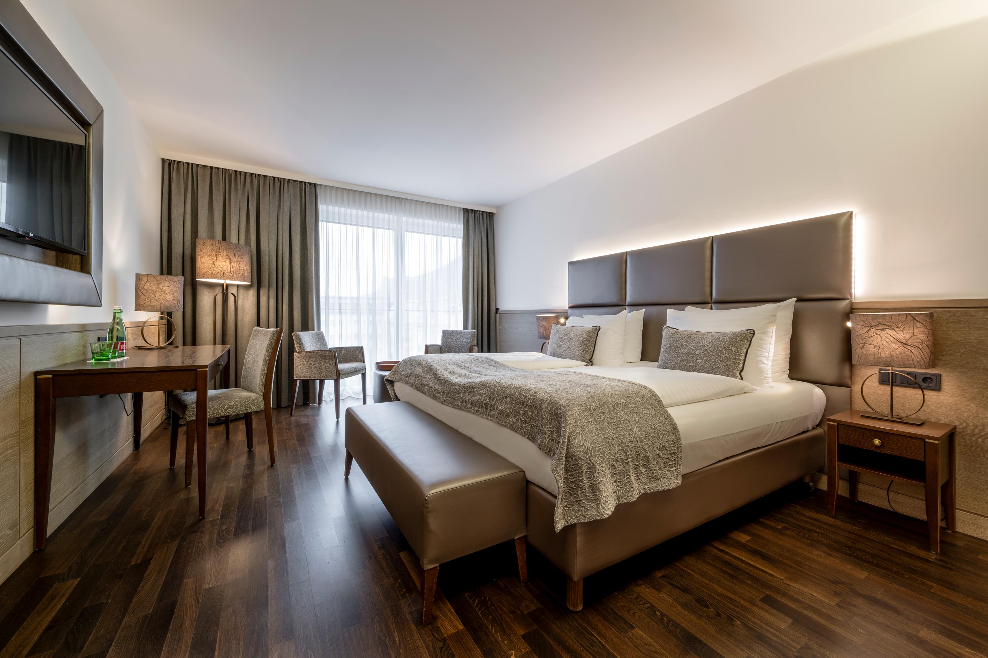 A modern hotel room with a large double bed and an elegant design. There is a desk and cozy seating as well as plenty of daylight.