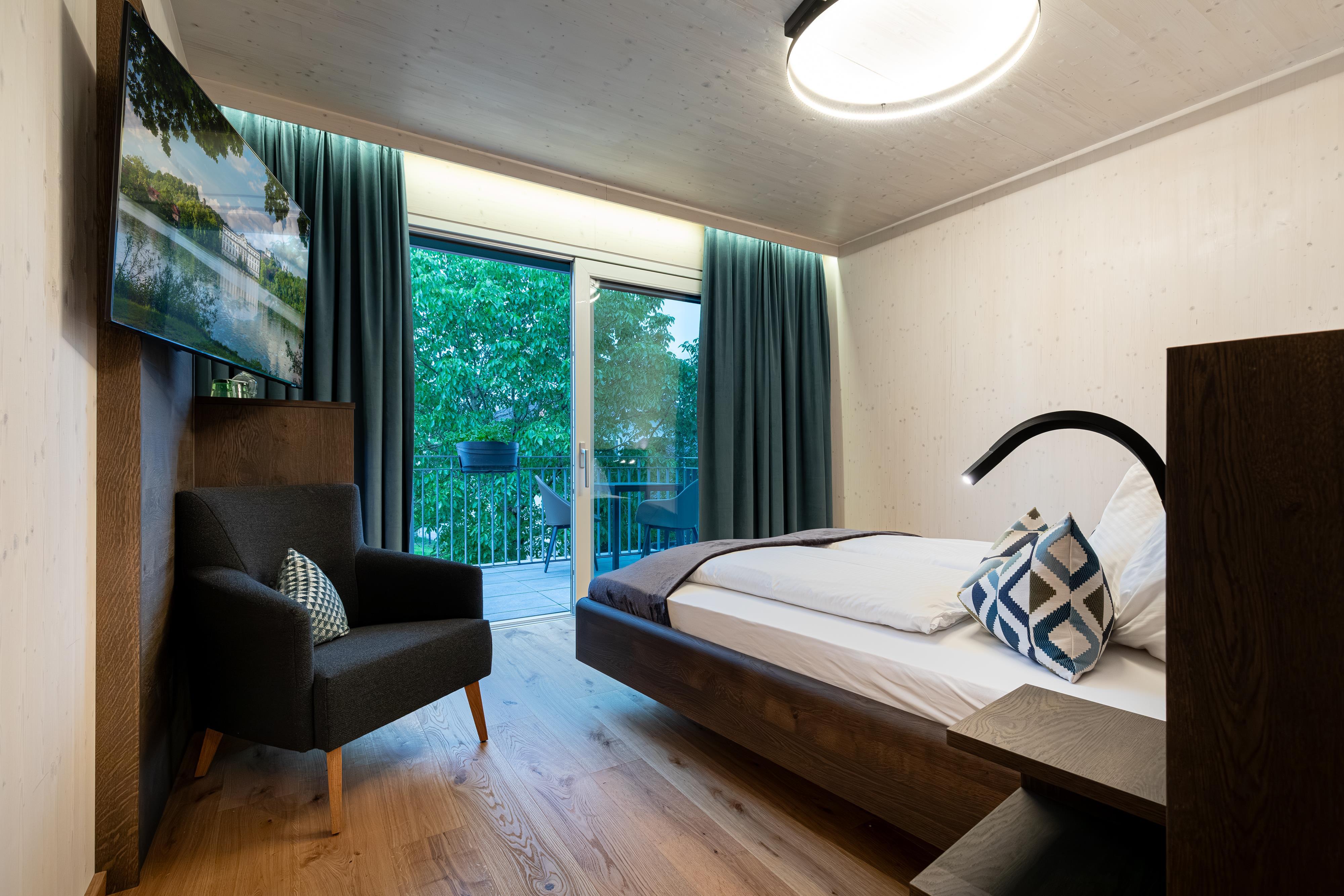 A modern bedroom with a double bed, a comfortable armchair, and a large window. The decor is simple and cozy with wood and green tones.