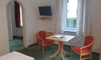 A simple room with two red upholstered chairs and a small table. A television hangs on the wall and a window provides daylight.