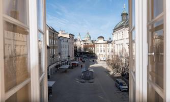 From a window, the view overlooks a quiet square with historic buildings. The sky is clear and blue, and there are trees and parked cars in the vicinity.