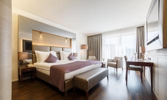 A modern hotel room with a large double bed and wooden floor. There are cozy seating areas and plenty of natural light.