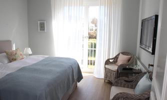 A bright bedroom with a large bed and soft colors. In front of a window, there is a cozy armchair and a TV on the wall.