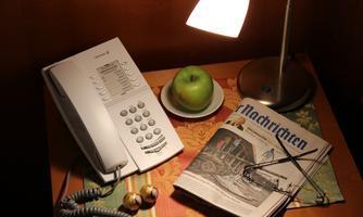 A table with a phone, a lamp, and an apple on a plate. Next to it lies a newspaper and a pair of glasses.