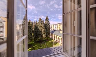 A view from an open window onto a green courtyard with tall trees. The sky is partly cloudy and the atmosphere is peaceful.