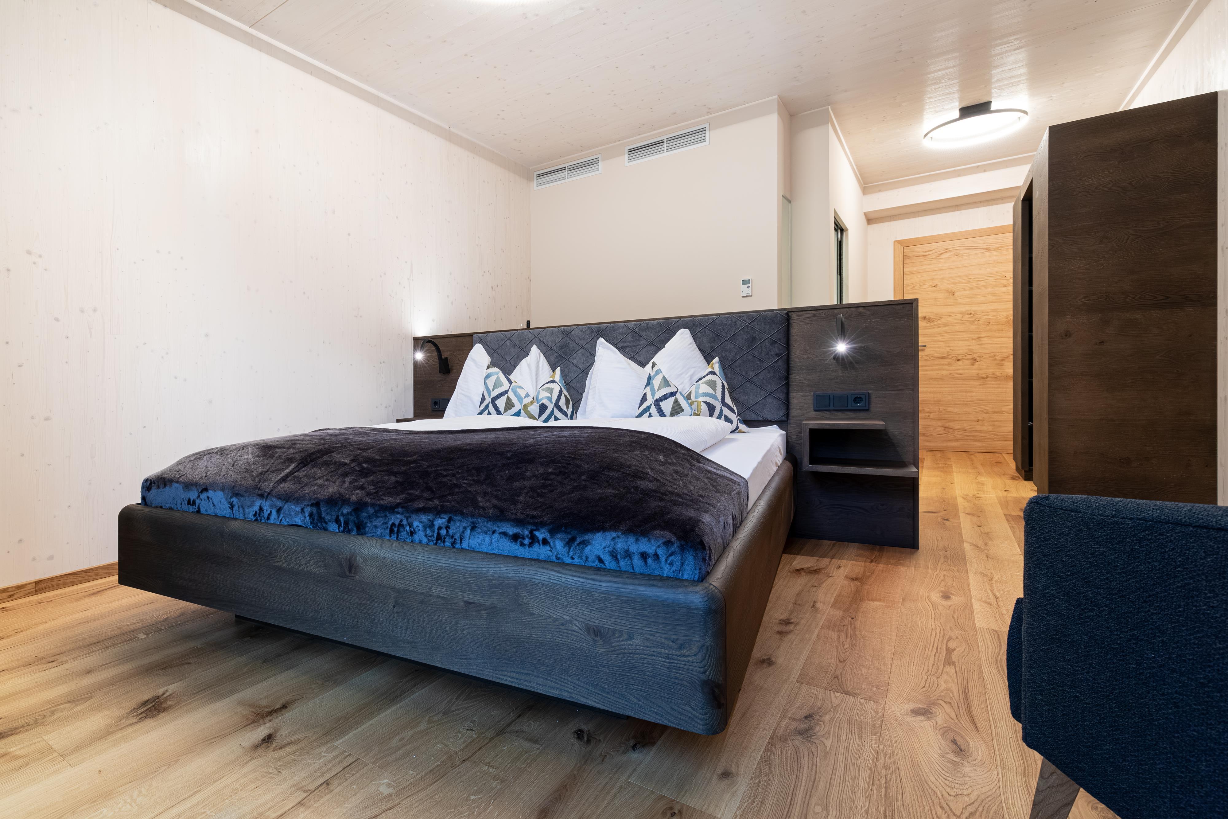 A modern bedroom with a large bed and cozy bedding. The room has wood-colored floors and an elegant, bright decor.
