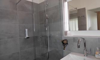 A modern bathroom with an elegant shower and a sink. The walls are designed with gray tiles and there is plenty of daylight.