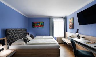 A modern room with two single beds and blue-painted walls. There is a desk, a television, and artwork that decorates the room.
