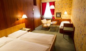 A cozy room with two beds, a table, and an armchair. The walls are decorated with a floral pattern and red curtains.