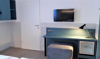 A modern hotel room with a television, a desk, and a chair. The walls are painted light and there is a closet.