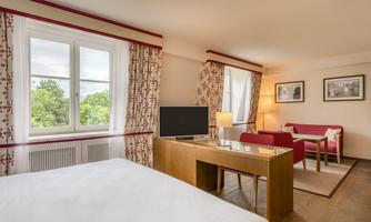 A bright hotel room with a comfortable bed, a table, and a television. Large windows provide plenty of natural light and a view of greenery.