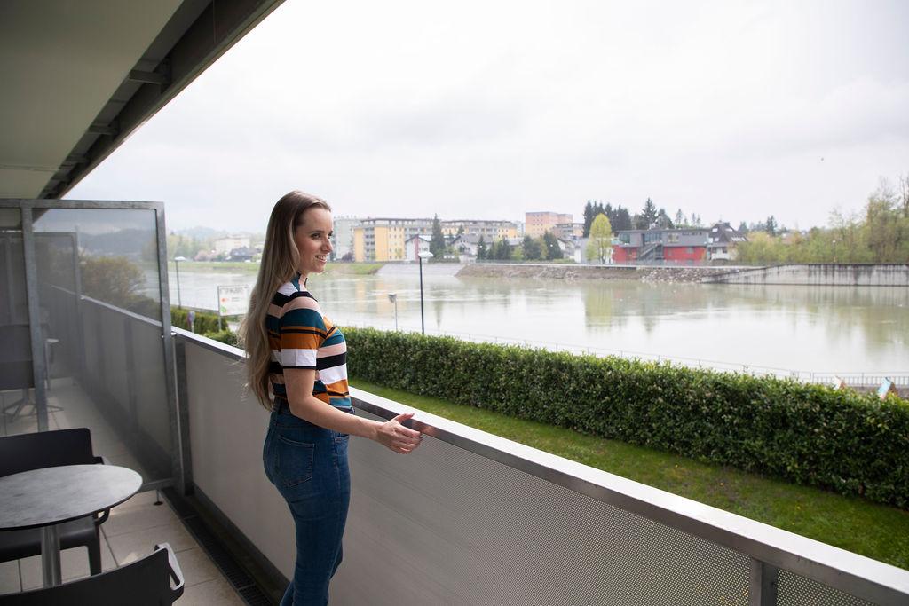 A woman stands on a balcony and looks at a river. In the background, buildings and trees can be seen, the sky is overcast.